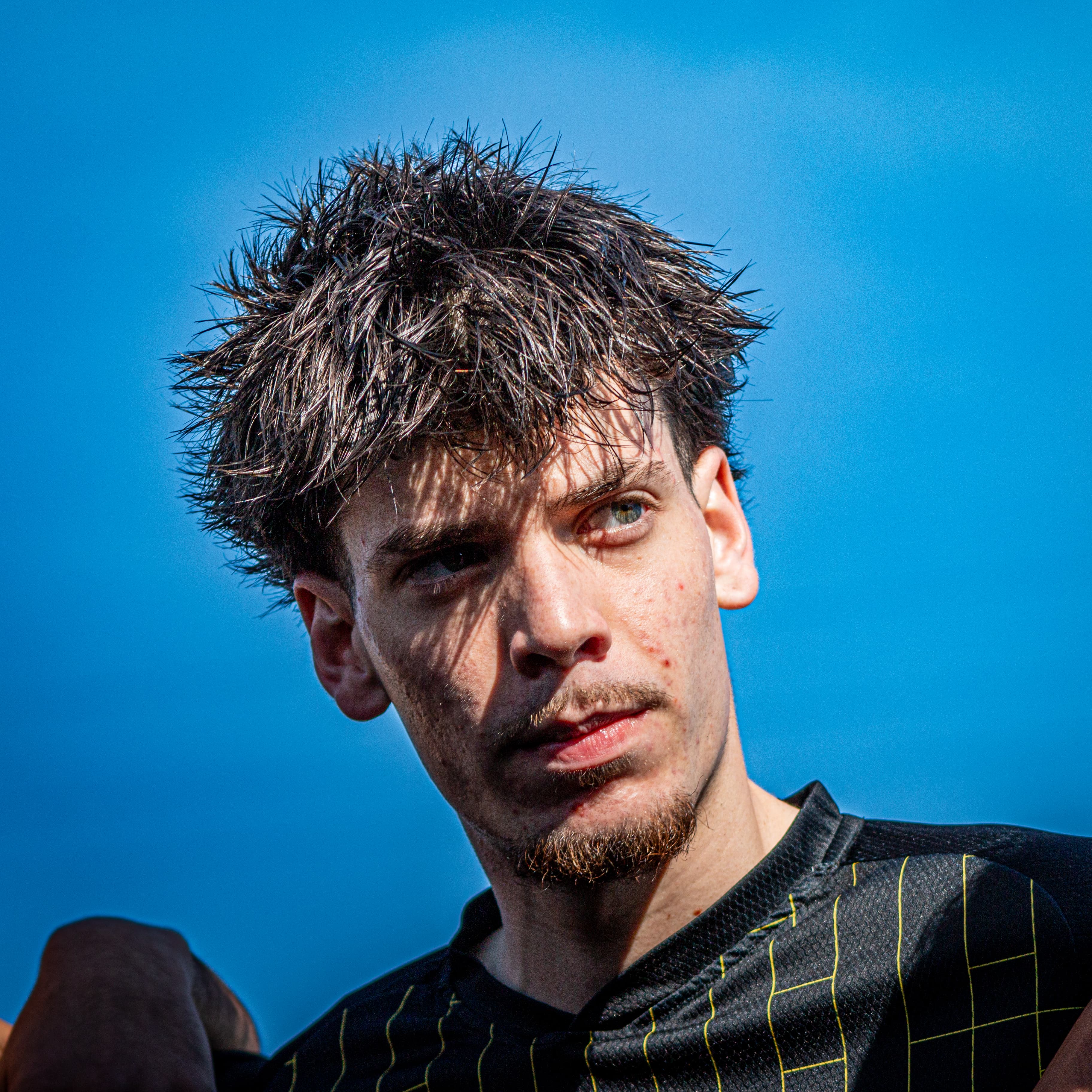Homem jovem com cabelo escuro bagunçado e barba curta, vestindo camisa preta sob céu azul.