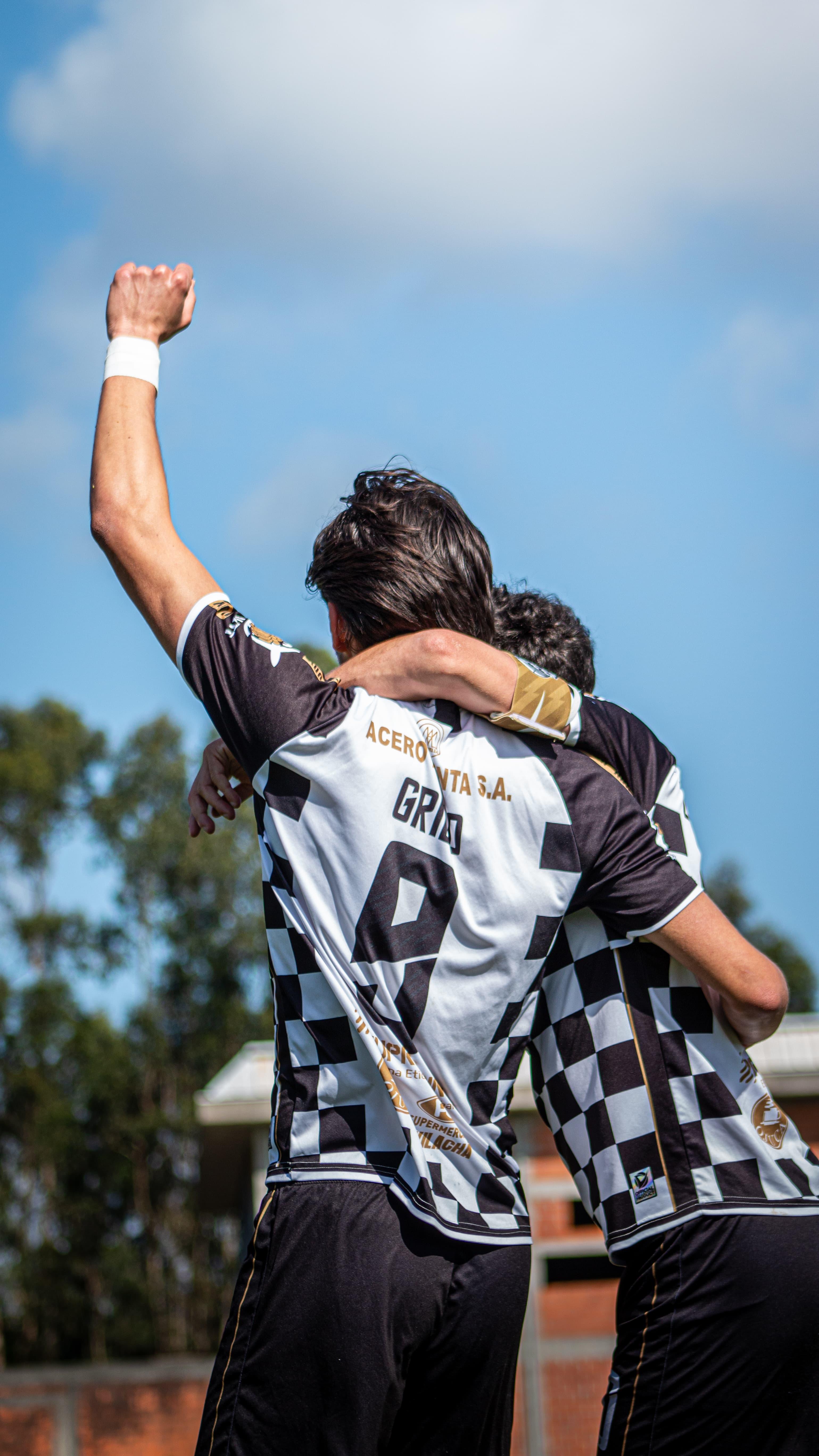 Dois jogadores de futebol com camisas quadriculadas pretas e brancas celebram com o punho erguido.