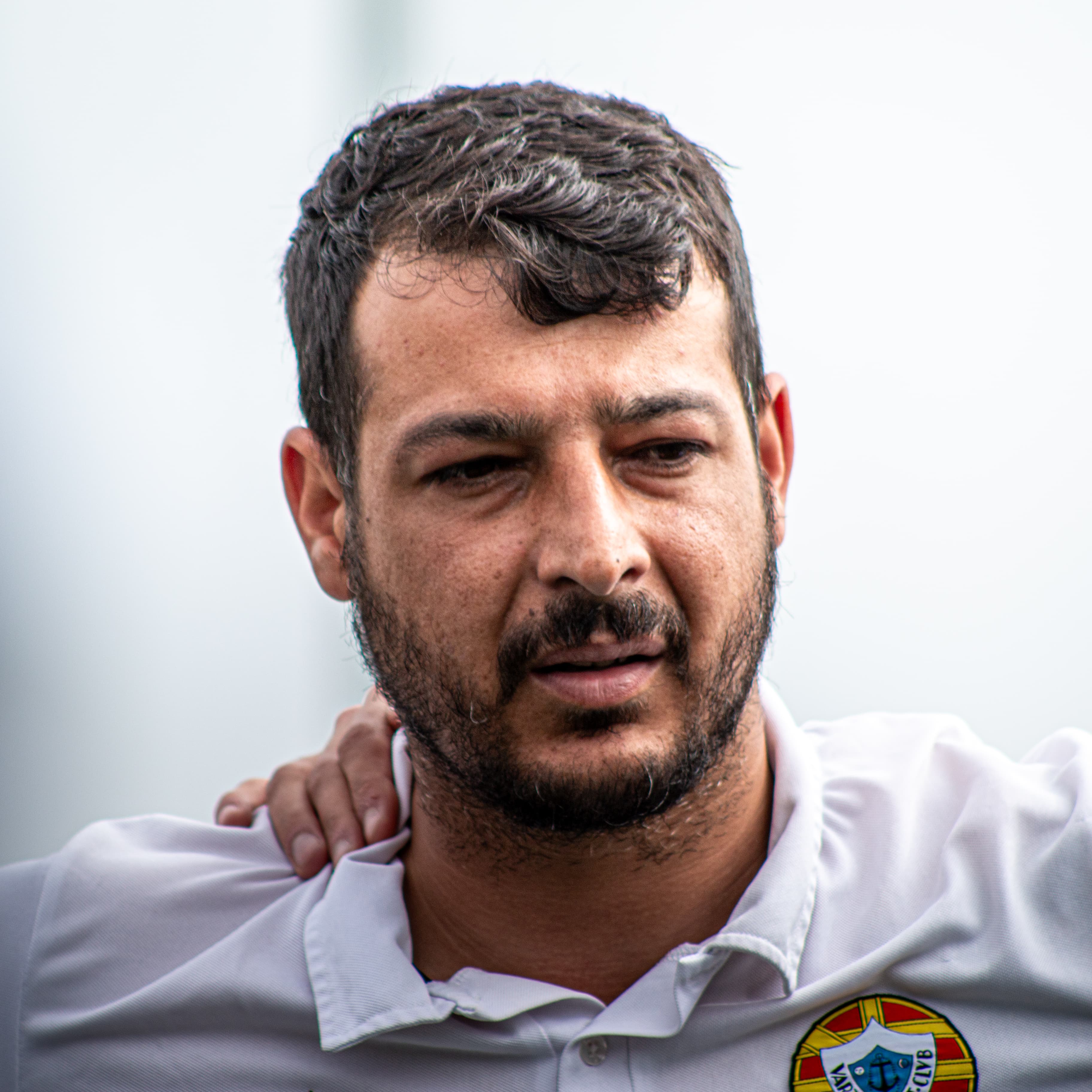 Homem com barba curta vestindo camisa polo branca com emblema e mão no ombro.