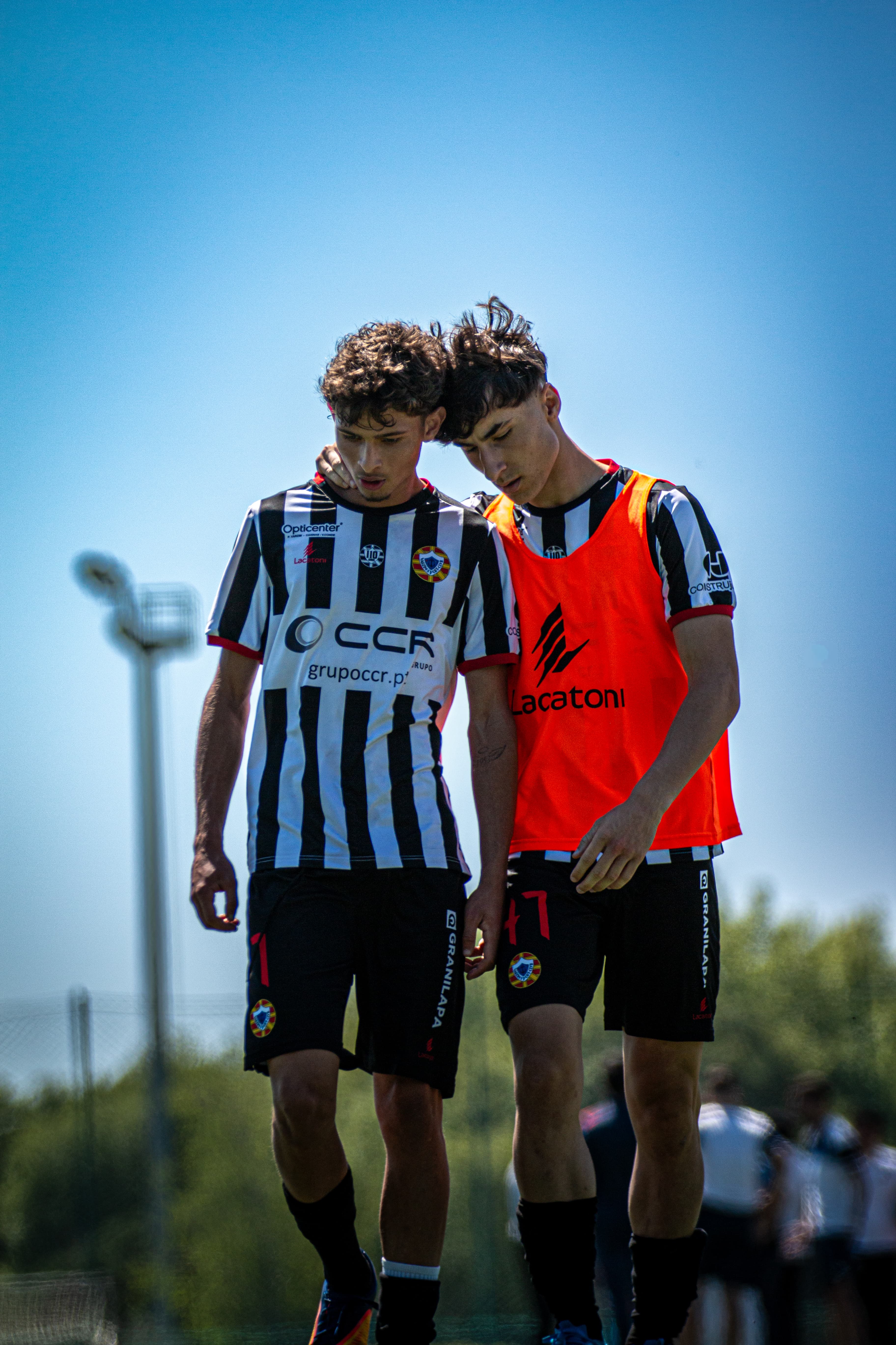 Dois jovens jogadores de futebol em uniformes listrados caminham juntos, um apoiando o outro.