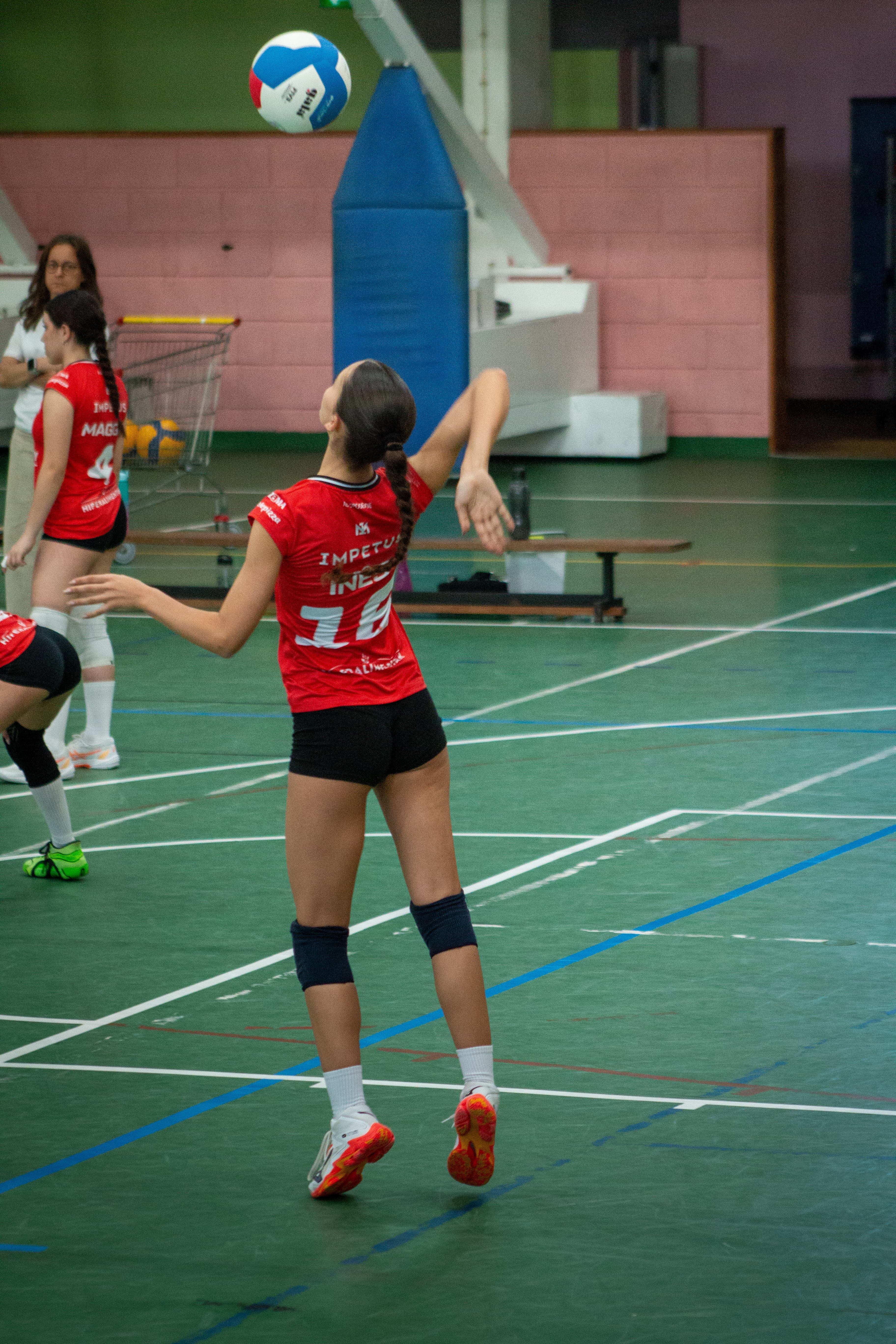 Jogadora de voleibol em uniforme vermelho salta para atingir a bola em quadra verde.