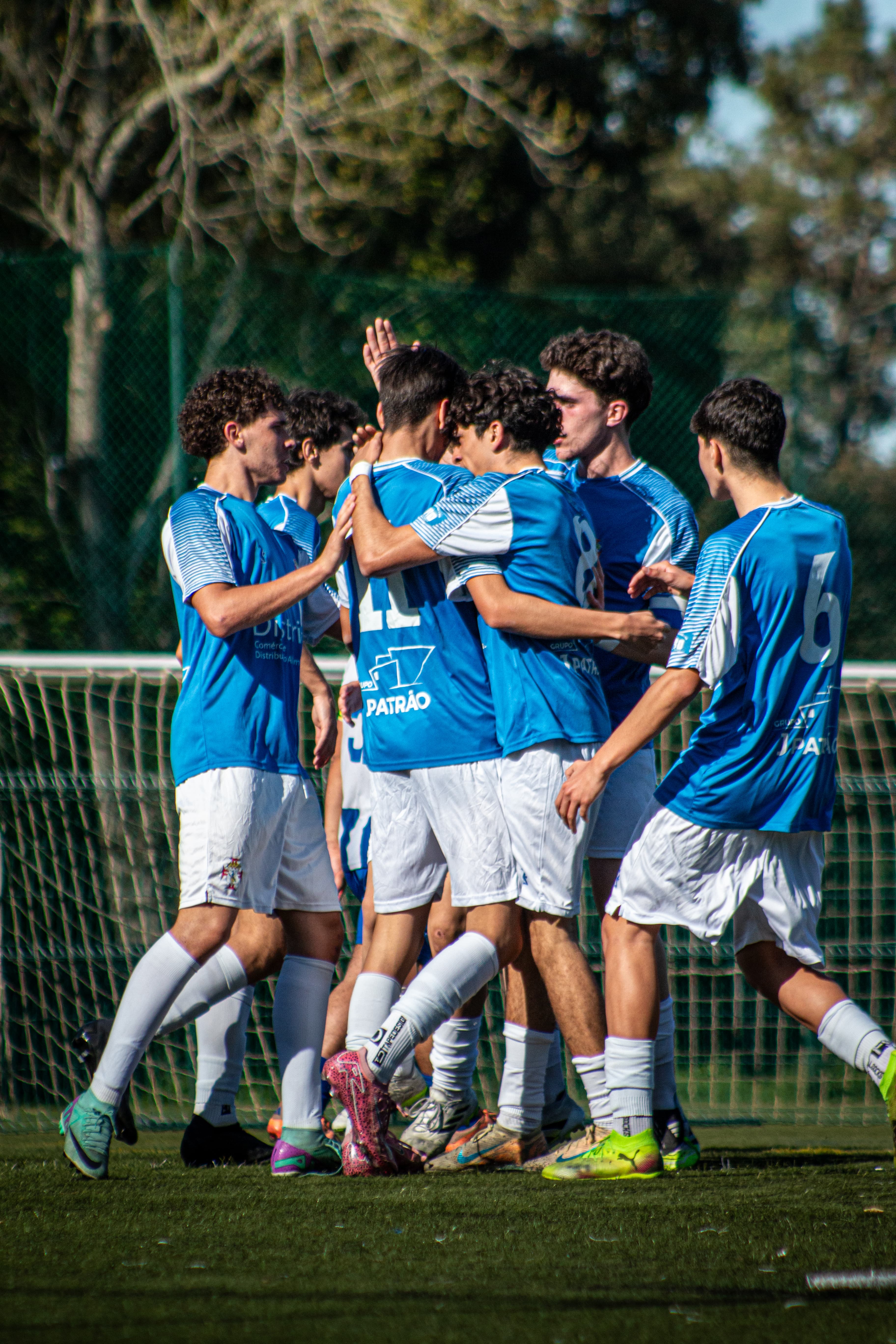 Jogadores de futebol jovens em uniformes azuis celebram um golo abraçados num campo ensolarado.