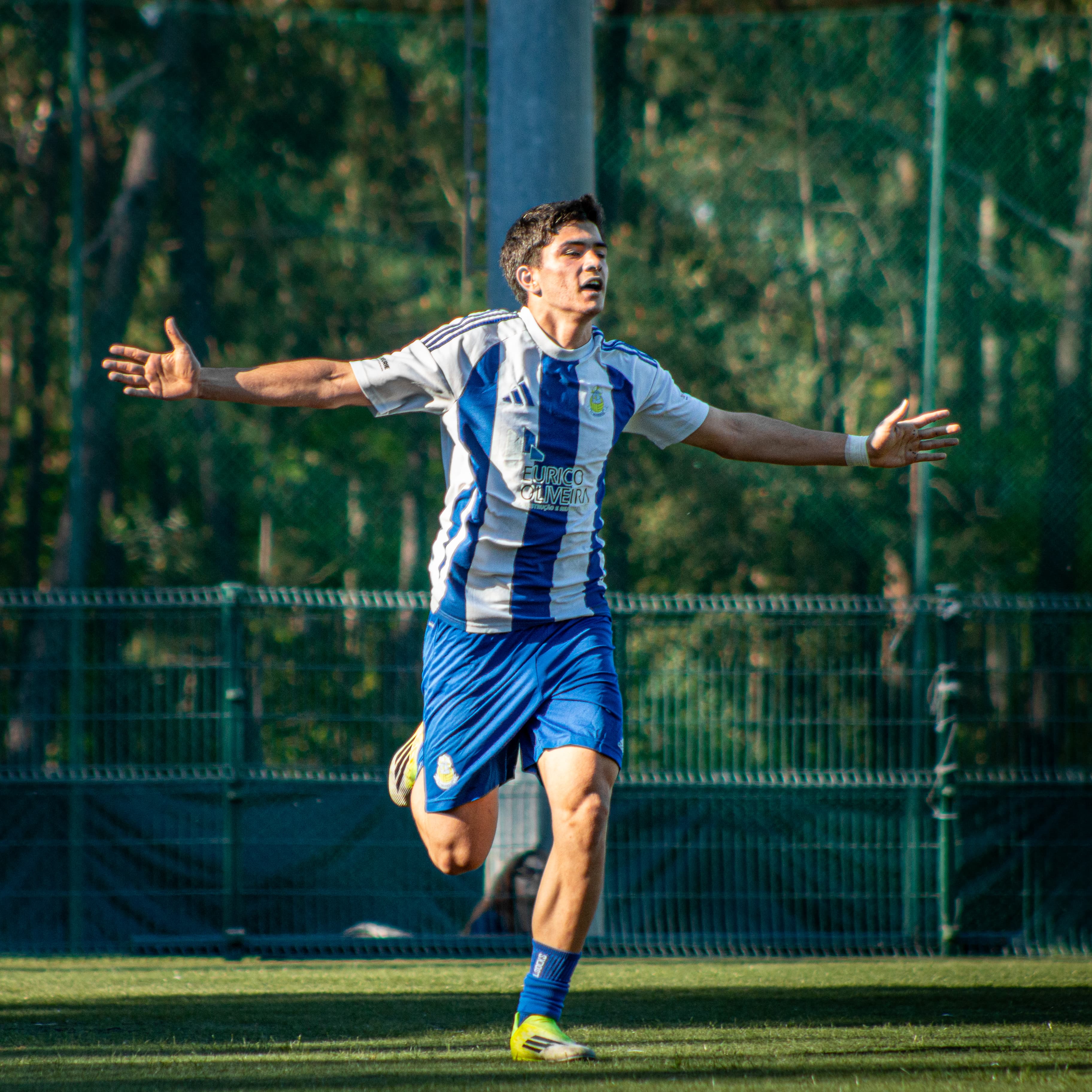 Jogador de futebol comemora com braços abertos, vestindo uniforme azul e branco em campo verde.