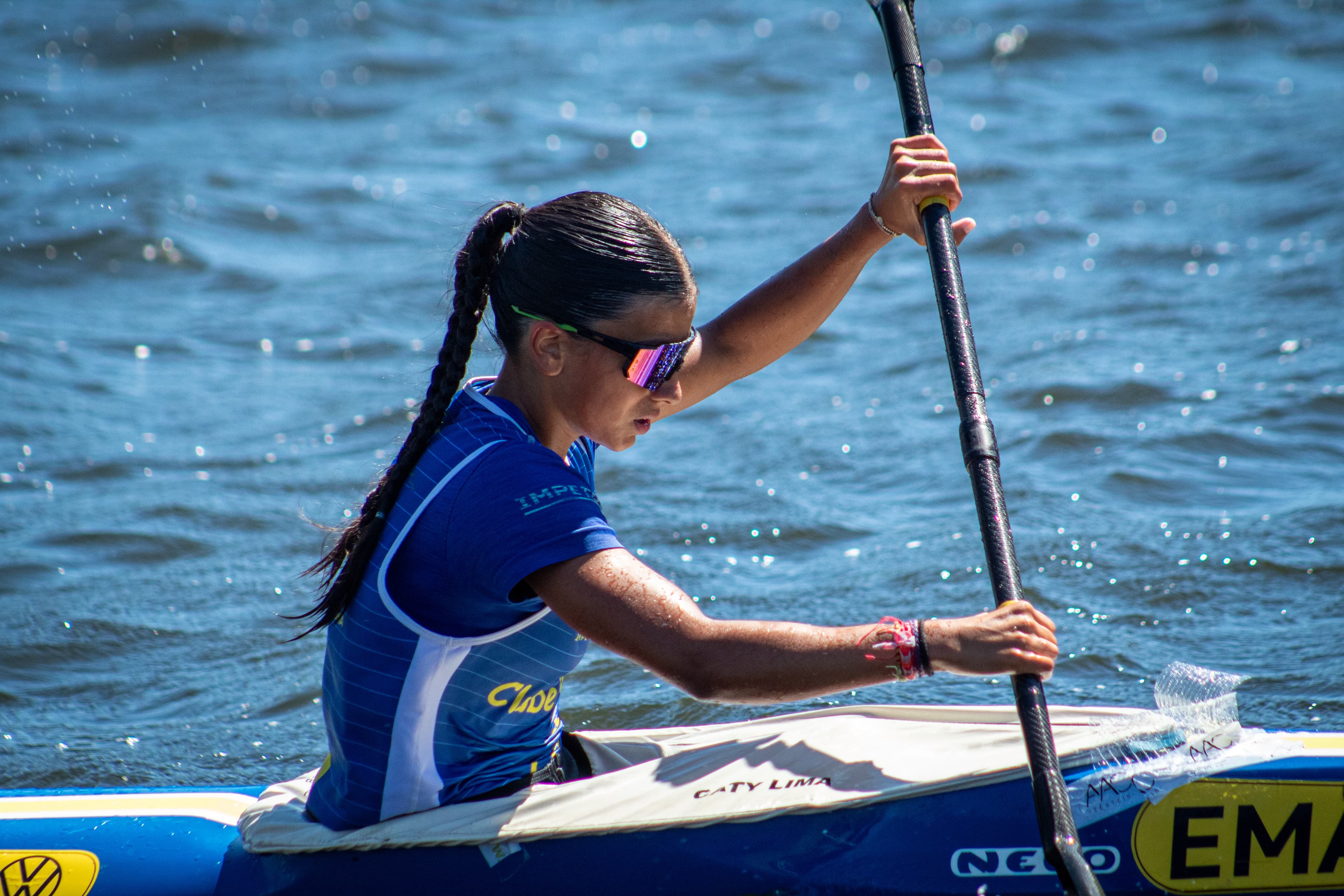 Atleta de canoagem com óculos escuros e trança rema caiaque azul em águas azuis brilhantes.