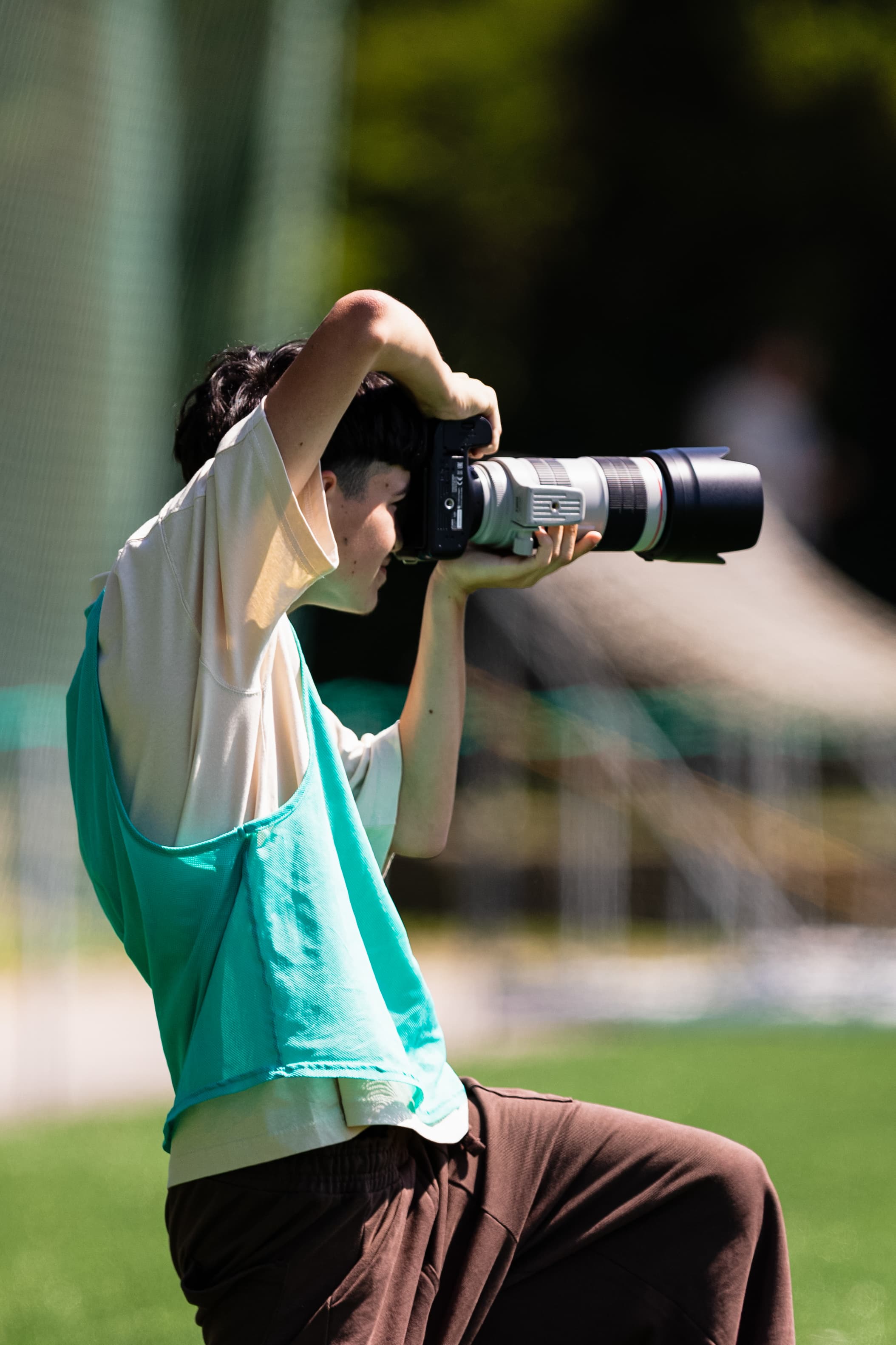 Fotógrafo jovem de colete verde segura câmera com lente teleobjetiva em um campo ensolarado.